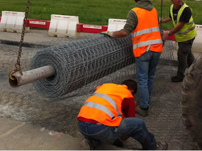 Three workers are spreading hexagonal wire mesh on a road, with the roll of mesh hanging on a circular tube, which is hung by a car.
