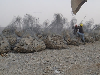 Gabion sacks are placed on a soil field, two men are hanging one of them onto a excavator.